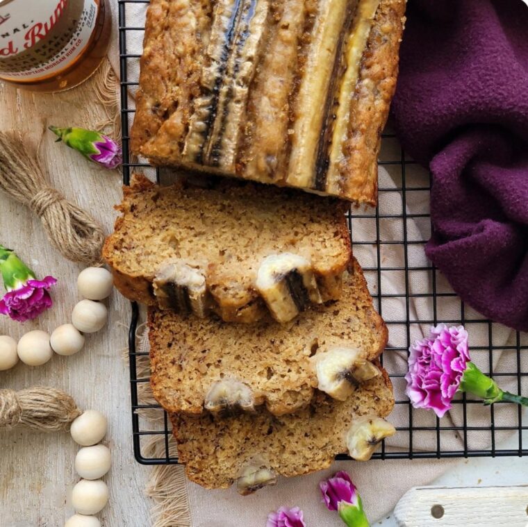 brown butter rum banana bread. top down view of a loaf on a black baking rack with three slices cut and resting on their side. image is styled with purple flowers and a bottle of captain morgan's rum.