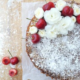 cherry buckle cake. top down view of uncut cake dusted with powdered sugar and garnished with tiny white carnations and stemmed cherries. background is wooden splattered with white paint and styled with 3 cherries in the bottom left