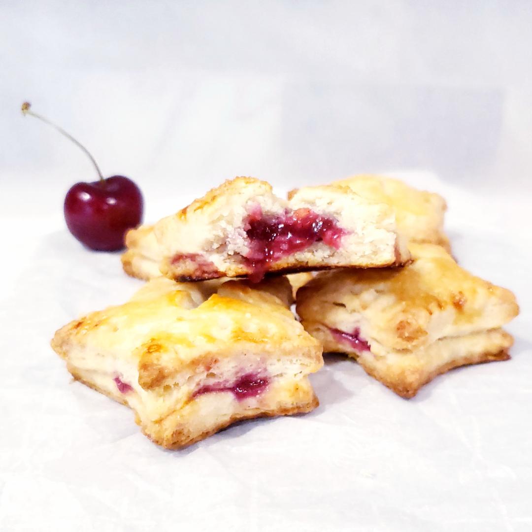 functional image cherry hand pies with pie crust star shaped black cherry in the background pies stacked on top of each other top pie has a bite out of it and is oozing cherry pie filling
