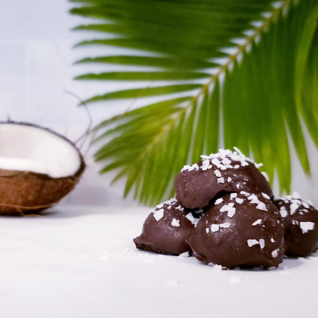 functional image dark chocolate coconut rum truffles stacked on a white counter with flaked coconut on top half a ripe cracked coconut in the background and a coconut palm frond in the background
