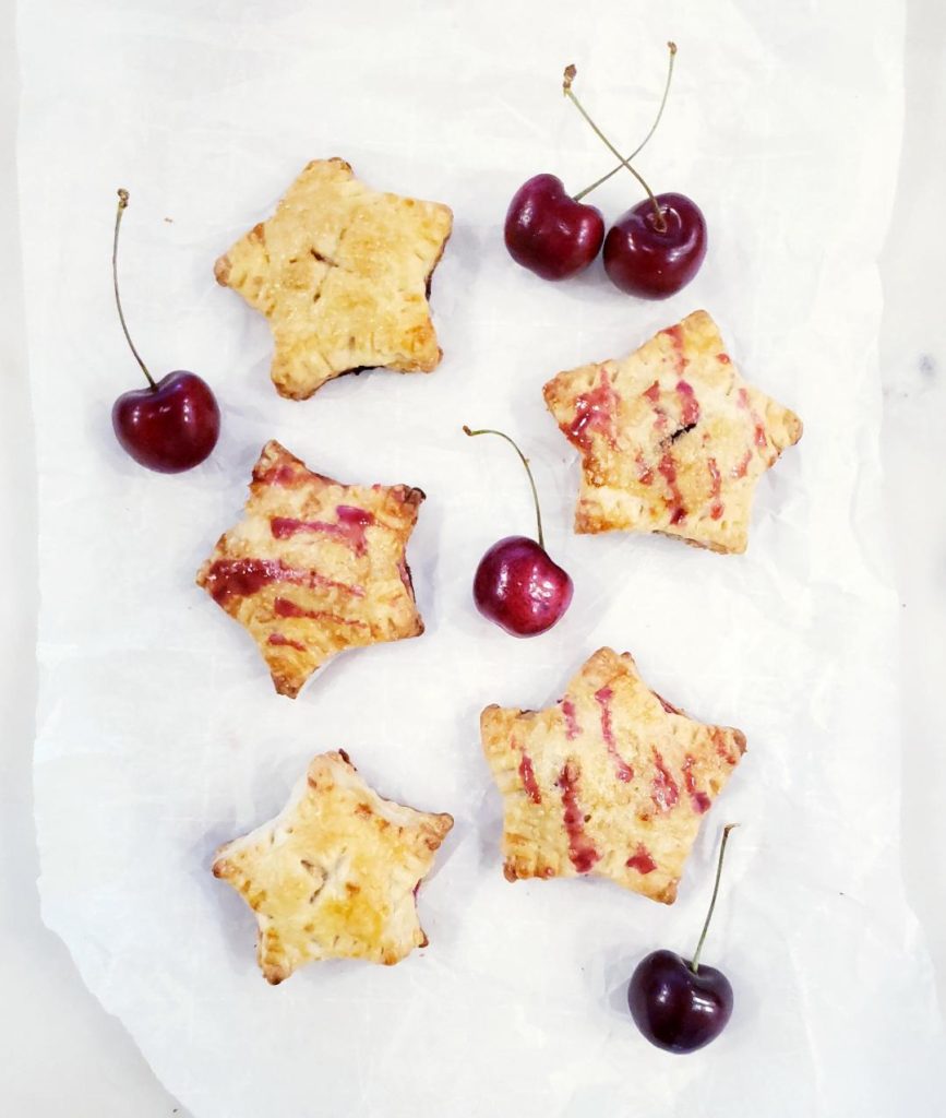 FUNCTIONAL image five star shaped cherry hand pies with pie crust on white parchment three cherry pies are drizzled with cherry juice food styled with fresh stemmed black bing cherries