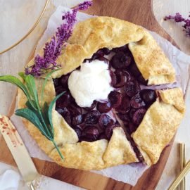 functional image black cherry galette top down with lavender a scoop of vanilla ice cream and one slice cut staged on a wooden cutting board with a knife covered in cherry juices