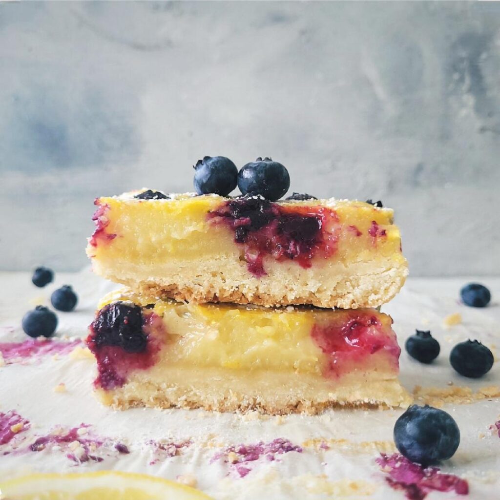 blueberry lemon shortbread bars. side view of 2 bars stacked on top of each other so you can see the layers of shortbread crust, lemon curd and blueberry topping.