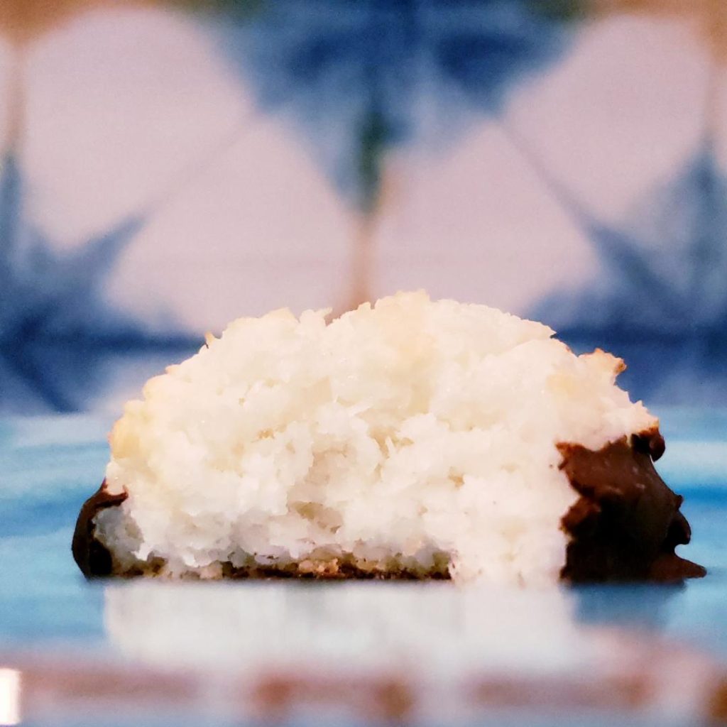functional image chocolate dipped coconut macaroon with a bite out of it so you can see the chewy moist coconut inside cookie is on a blue plate blue and has a  white geometric background 