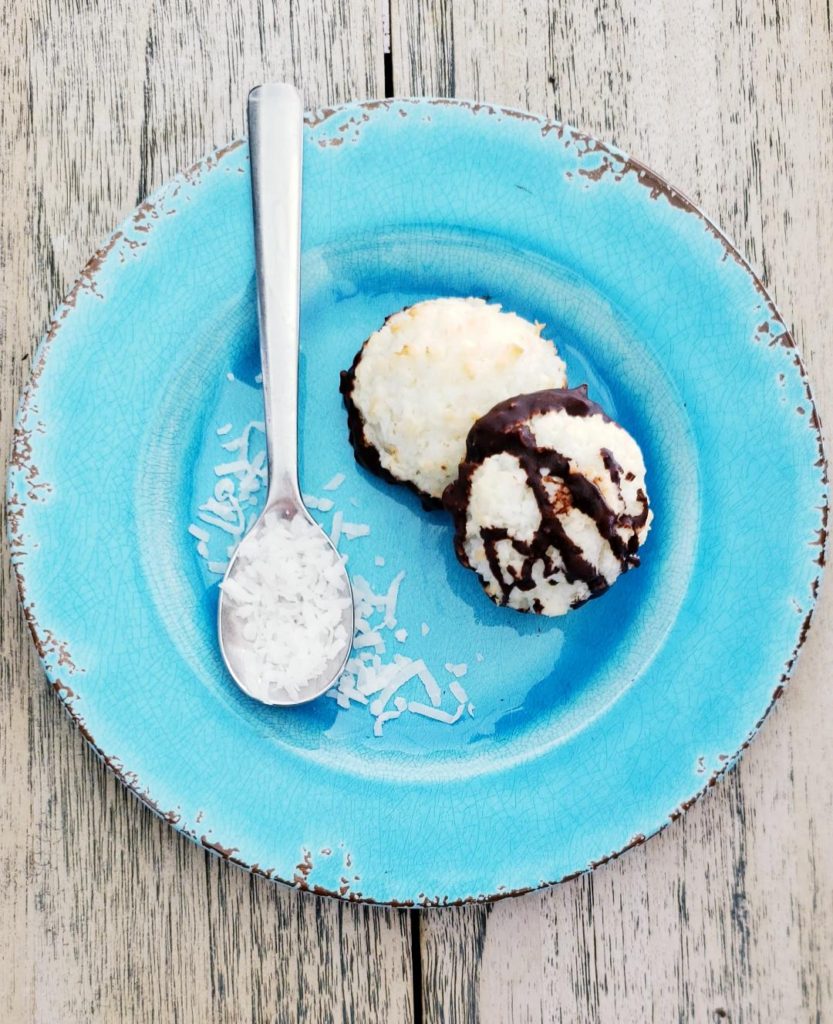 functional image distressed and cracking finish on blue plate with two coconut macaroons one white and bottom dipped in dark chocolate the other dipped and drizzled in dark chocolate silver spoon on plate with coconut flakes on it plate is on washed out wood background chocolate dipped coconut macaroons