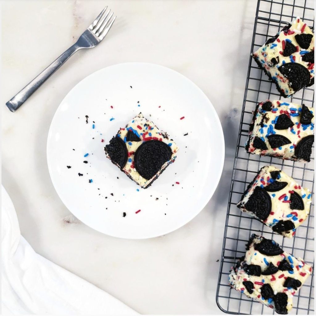 functional image oreo funfetti cheesecake bars one square slice on a white plate fork in upper left corner four square slices running down the right side on a cooling baking rack