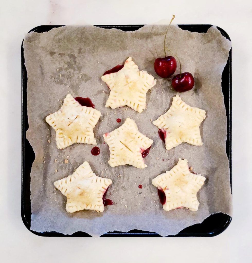 functional image six star shaped cherry hand pies with pie crust on a square black baking sheet with parchment paper and two fresh cherries with stems hand pies are oozing cherry filling, edges crimped with fork and pies are ready to be baked in the oven