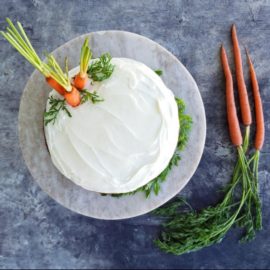 functional image carrot cake bourbon cream cheese frosting top down view distressed gray blue table cake stand is a light tan marble three carrots with stems to the right three carrot tops inserted in top of cake