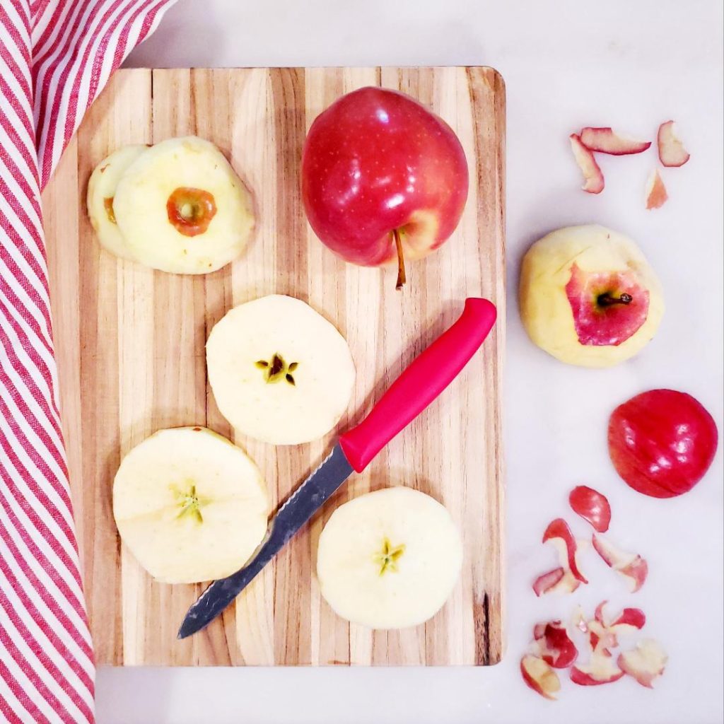 functional image bourbon apple crisp preparation top down photo of a wooden cutting board with cut apples and a red handled knife red and white striped linen apple peels and a red apple tilted on its side skillet apple crisp