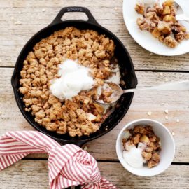 functional image bourbon apple crisp in a cast iron skillet top down photo on a distressed wood plank background skillet handle is wrapped with a red striped linen three scoops of vanilla ice cream on top of crisp one bowl and one plate on the right side with servings of crisp and ice cream