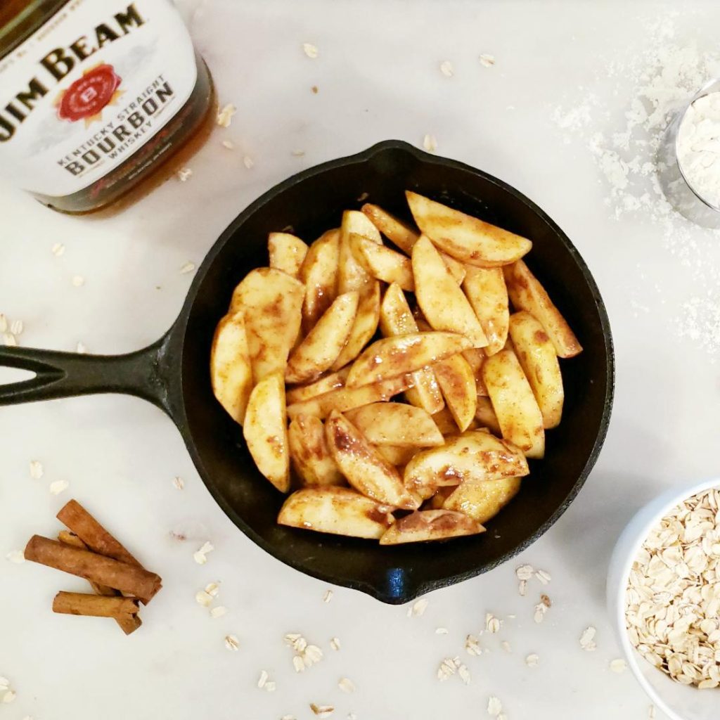 functional image bourbon apple crisp top down photo on white marble cast iron skillet filled with sliced apples coated with cinnamon sugar bottle of  jim beam bourbon in upper left corner cinnamon sticks in lower left corner measuring cup of flour in upper right corner and bowl of oats in lower right corner skillet apple crisp