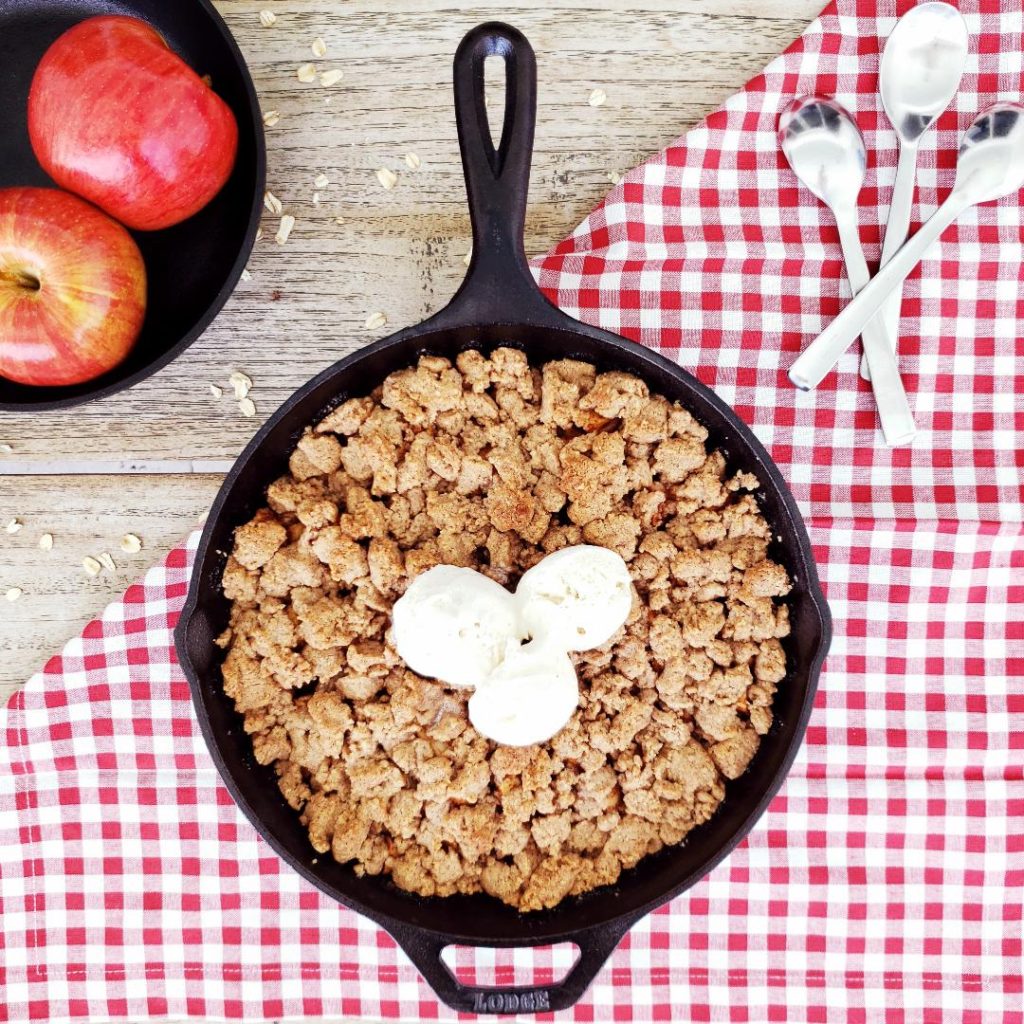 functional image top down view of bourbon apple crisp in cast iron skillet with three scoops of vanilla ice cream on a distressed wooden surface with a gingham red linen and three spoons in upper right corner upper left corner has two red apples in a smaller black skillet apple skillet crisp