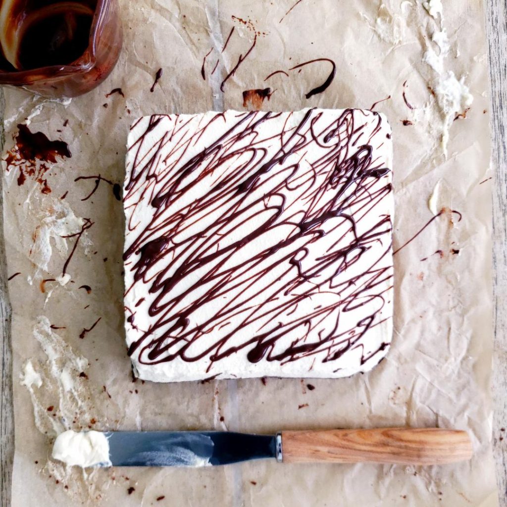 baileys brownies top down view of an 8x8 pan of brownies uncut and frosted with baileys frosting and drizzled with melted chocolate. surface is messy brown parchment with lots of drips of chocolate and smears of frosting