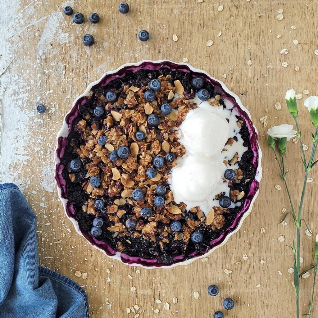 blueberry almond crisp in a white ceramic baking dish topped with fresh blueberries and two scoops of melting vanilla ice cream. top down view of crisp. background is wooden and is styled with tiny white carnations, a denim blue linen, oats and fresh blueberries