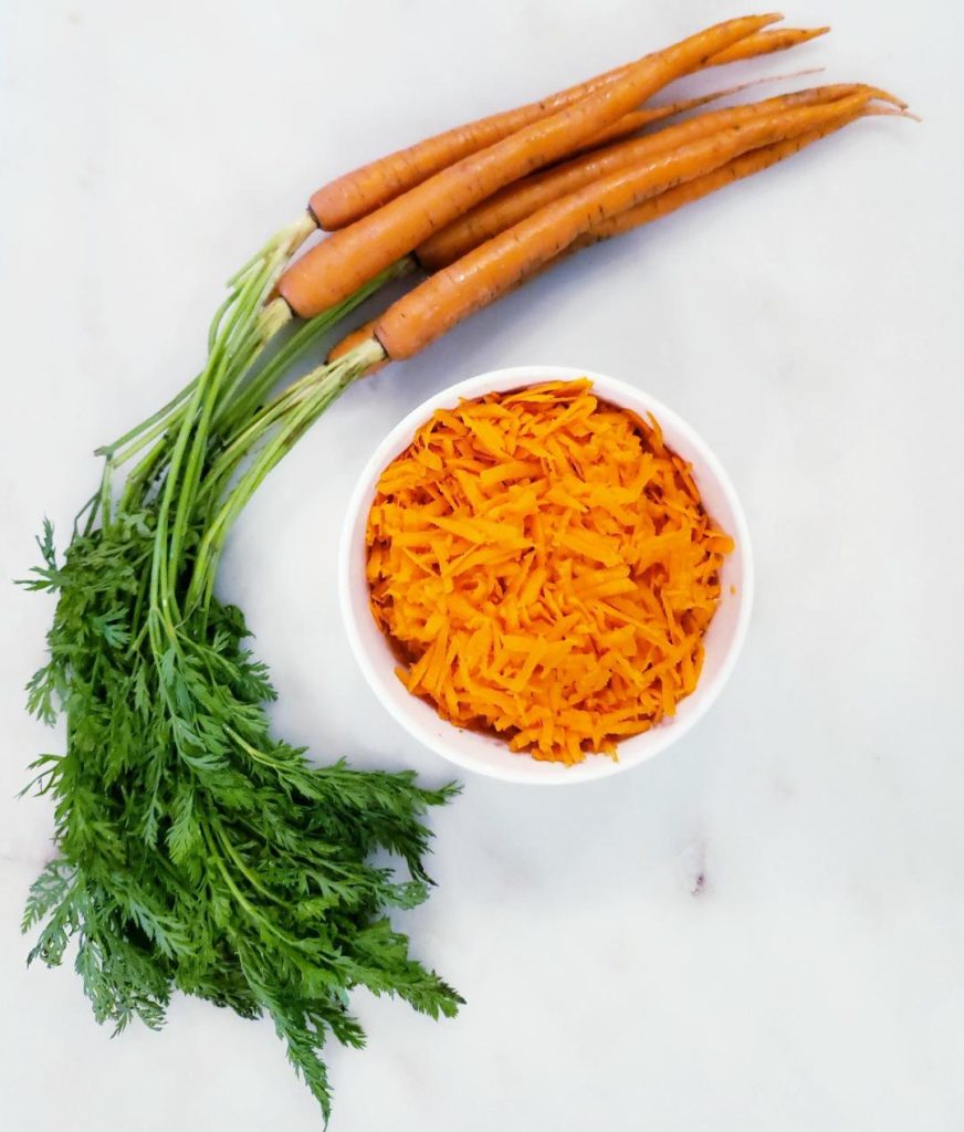 white bowl of shredded carrots on white marble with freshly picked carrots with stems still on surrounding bowl on left side carrot cake with bourbon cream cheese