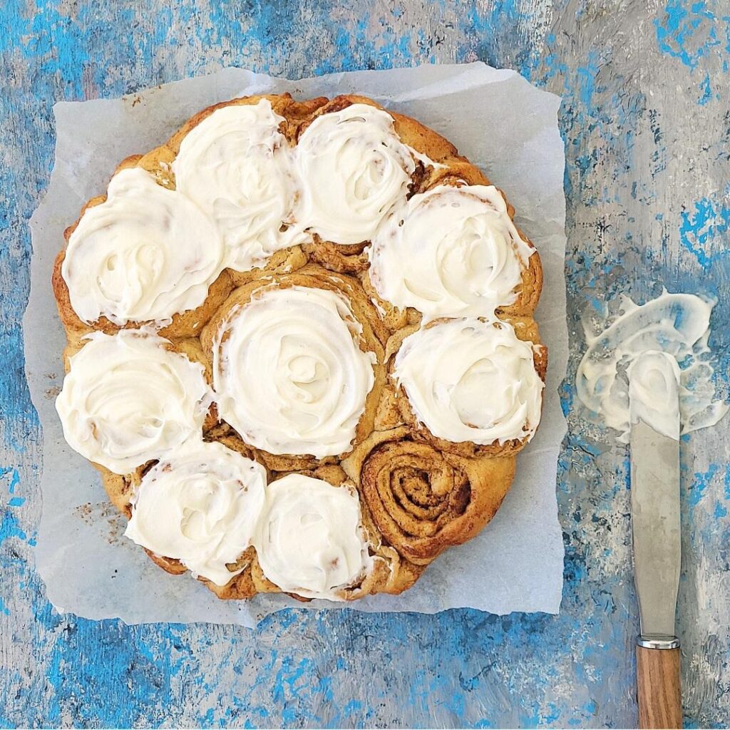 soft and fluffy cinnamon rolls with cream cheese frosting. top down view of rolls all but one are covered in icing. background is blue and there is a metal spatula to the left with cream cheese on it.