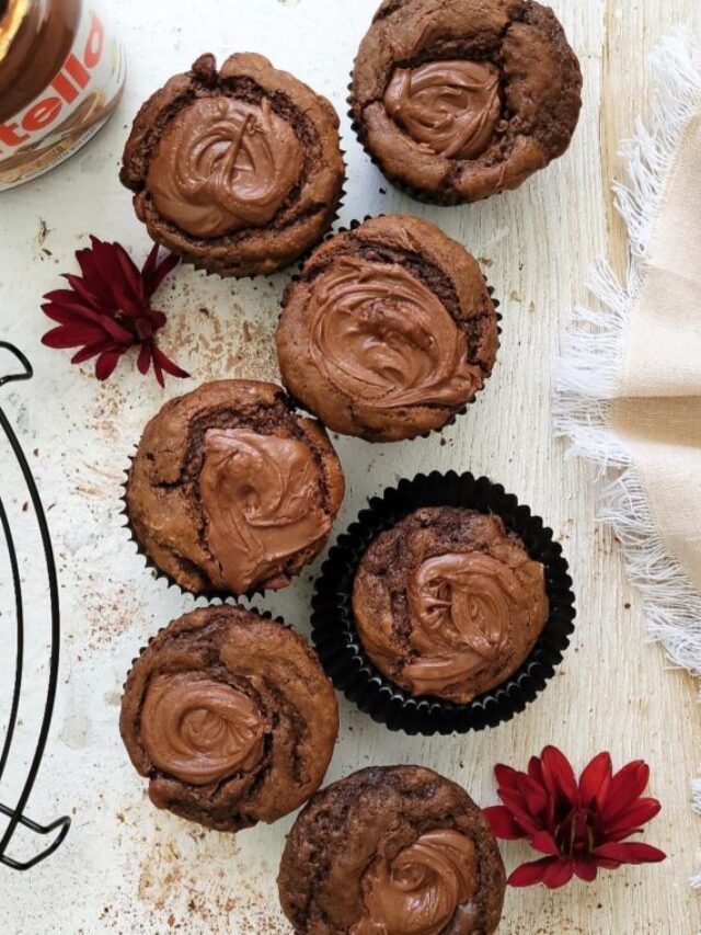chocolate nutella muffins. top down view of 7 muffins on a white wood surface. image is styled with a round wire baking rack, a jar of nutella, maroon fall flowers and a fringed linen. tops of muffins are swirled with nutella.