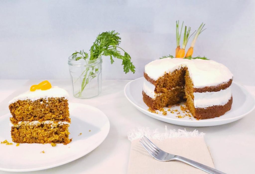 carrot cake iced with cream cheese frosting on a large white plate one piece is cut and on a smaller white plate fork and fringe linen napkin in foreground and carrot stems in a glass in the background carrot cake with bourbon cream cheese