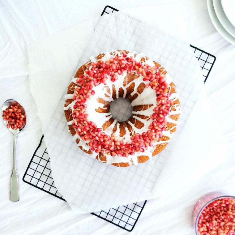 functional image bourbon honey bundt cake top down bright white glade and red pomegranate seeds as decoration cake sits on white parchment paper on top of a black baking rack styled with a spoon full of pomegranate seeds and a bowl full of pomegranate seeds