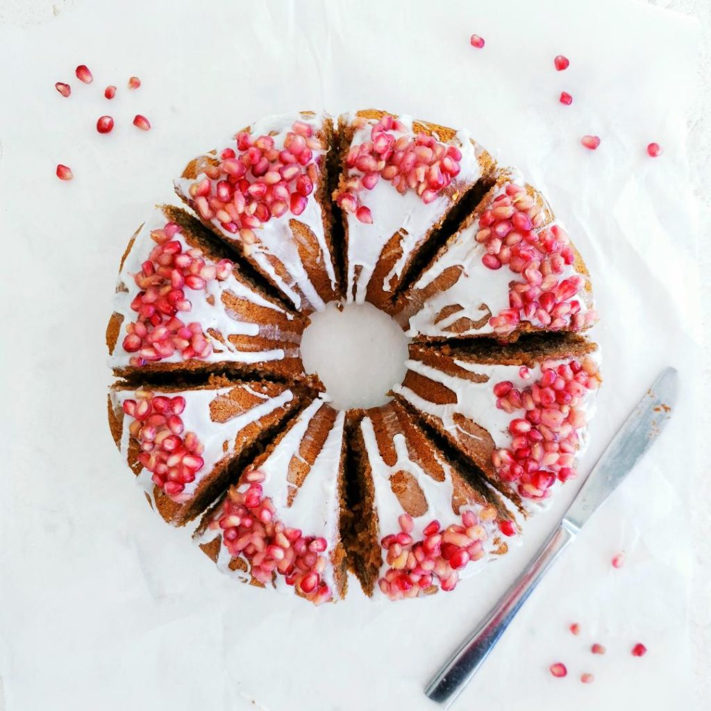 functional image bourbon honey bundt cake top down cut into 8 giant slices with white glaze and pomegranate seeds as decoration on white background with a stainless steel knife