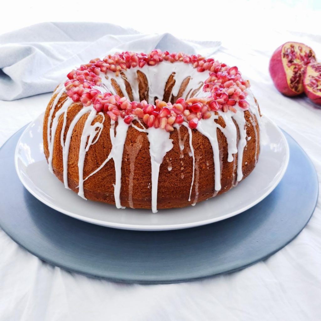 functional image baked bourbon honey bundt cake with vanilla glaze and pomegranate seeds garnish side view on white plate on a gray charger cut pomegranate in the back right gray linen in back left background is white