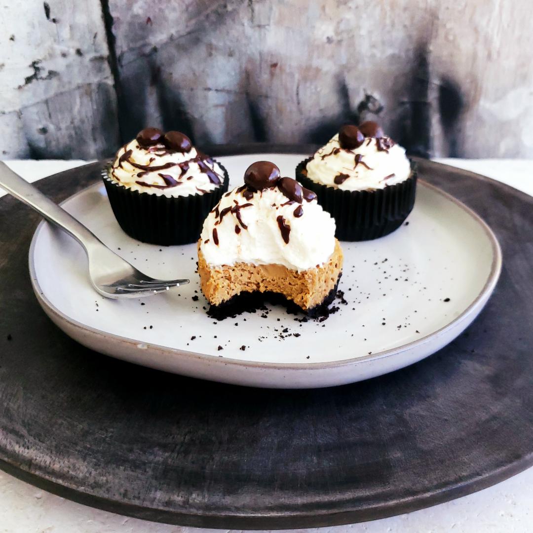 functional image three mini baileys mocha cheesecakes on a light gray plate on top of a dark gray charger one mini cheesecake in front and two in back front cheesecake has a bite out of it so you can see the contrasting layers of black oreo cookie crust coffee colored cheesecake and white whipped cream