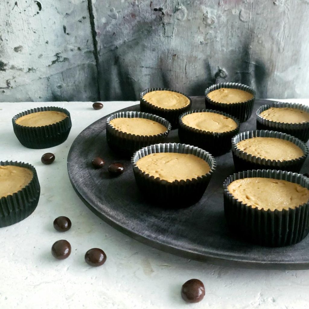 functional image mini baileys mocha cheesecake in black cupcake wrappers fresh from the oven no topping applied yet side view distressed gray wooden plate with 8 randomly placed cheesecakes and two additional cheesecakes to the right on white marble styled with chocolate covered espresso beans background is abstract distressed wood painted gray white black 