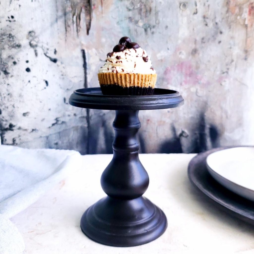 functional image mini baileys mocha cheesecake side view one mini cheesecake sitting on top of a black candlestick white counter abstract painted background gray white black white plate and gray charger to the right and a gray linen to the left