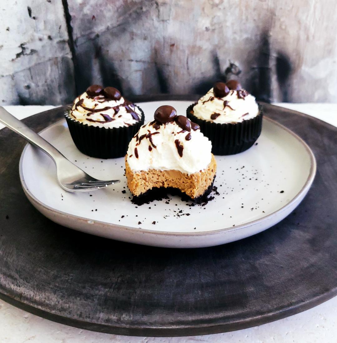 functional image three mini baileys mocha cheesecakes on a light gray plate on top of a dark gray charger one mini cheesecake in front and two in back front cheesecake has a bite out of it so you can see the contrasting layers of black oreo cookie crust coffee colored cheesecake and white whipped cream