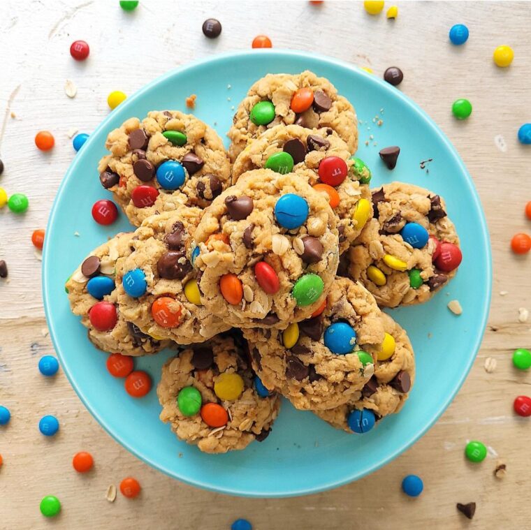 easy monster cookies. m&m cookies top down view of cookies piled on a pale blue plate. background is scattered with colorful m&m's
