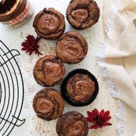 chocolate nutella muffins. top down view of 7 muffins on a white wood surface. image is styled with a round wire baking rack, a jar of nutella, maroon fall flowers and a fringed linen. tops of muffins are swirled with nutella.
