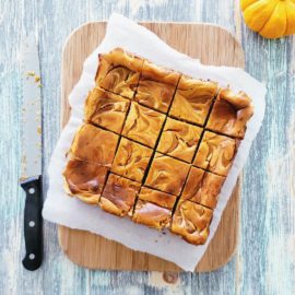 functional image pumpkin swirl cheesecake bars out of the oven and cut into 16 squares on white parchment paper and a wooden cutting board top down photo dirty blade knife with black handle on left and mini pumpkin in upper right background is distressed light blue wood