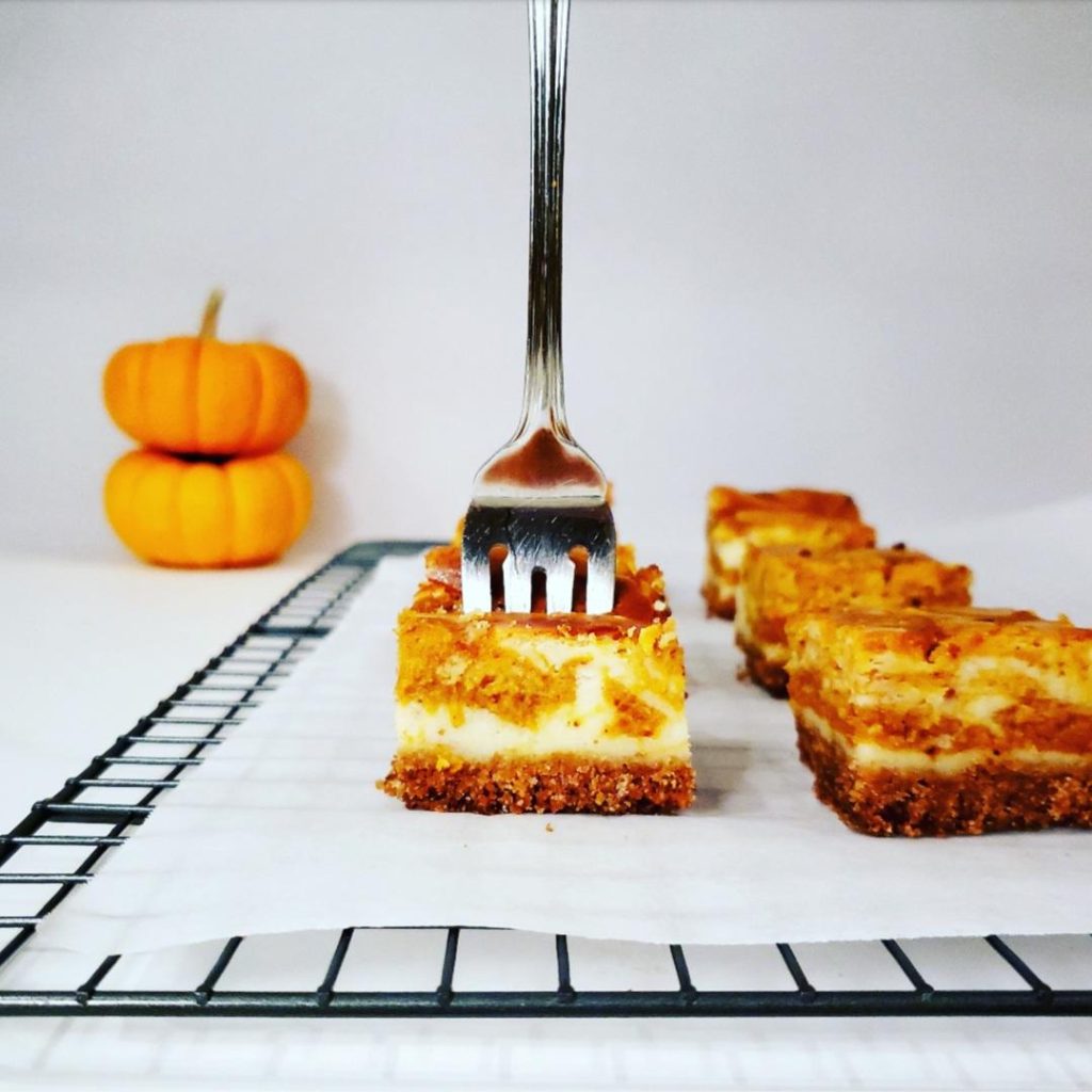 pumpkin cheesecake bars with graham cracker crust cut into squares side view photo of slices on white parchment paper on top of a black cooling rack front left square has a fork through the top two mini pumpkins stacked in the background