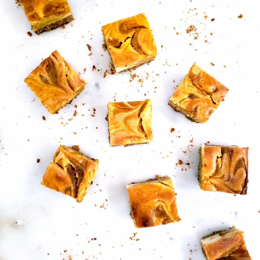 pumpkin cheesecake bars with graham cracker crust cut into squares and placed randomly on a white marble counter top down photo with crumbs scattered