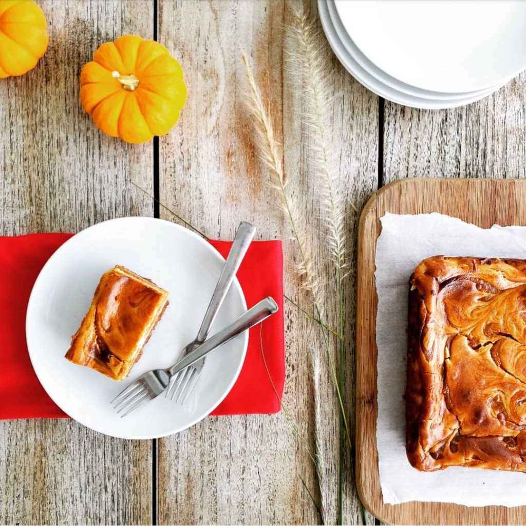 functional image cheesecake bars pumpkin pumpkin swirl cheesecake bars graham cracker crust on distressed wooden planks with two mini pumpkins in upper left corner white plates stacked in upper right corner styled with wheat grass in the middle red linen folded under a white plate on the left with two forks and a rectangular slice of pumpkin swirl cheesecake bars top down view lower right of photo is a wooden cutting board with white parchment and an uncut batch of pumpkin swirl cheesecake bars