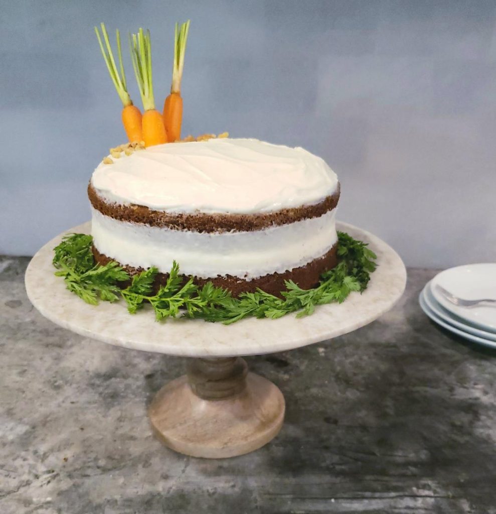  bourbon cream cheese frosted layer cake carrot cake on a wooden cake stand encircled by green carrot leaves decorated with peeled carrot tops on crown of cake white plates stacked in background with a fork carrot cake with bourbon cream cheese 