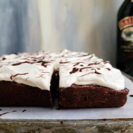 baileys brownies with baileys whipped cream side view of frosted brownies with a bottle of baileys in the back right background is blue and gray abstract