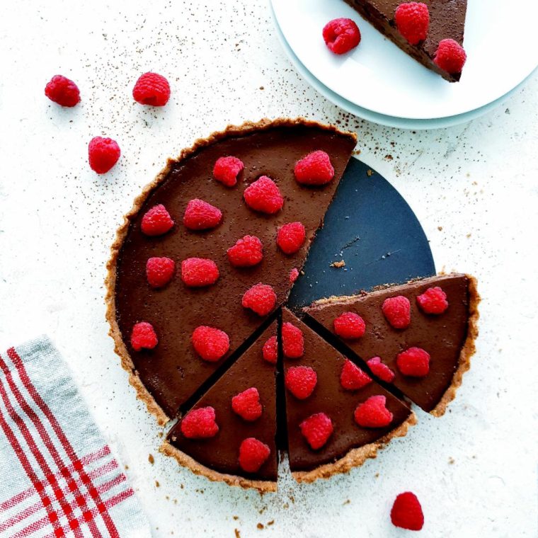 functional image dark chocolate raspberry tart top down photo on white limestone chocolate tart one half not sliced the other half is sliced one slice is on a white plate in upper right corner other slices are still on the tart pan bottom left bottom corner is a red striped lined tart photo is styled with a few loose raspberries