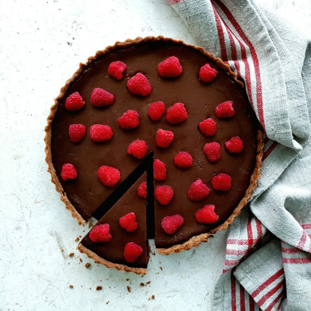 functional image raspberry chocolate tart top down photo one slice has been cut and slightly removed a red striped linen is to the left and there are a few crumbs surrounding the cut slice