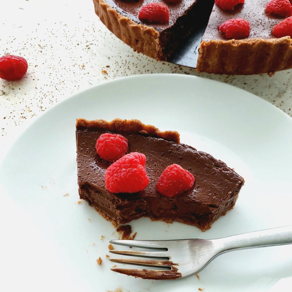 functional image raspberry chocolate tart top down image one slice of tart on a white plate with a chocolaty fork tart slice has three raspberries on top. in top right corner is the tart minus the slice