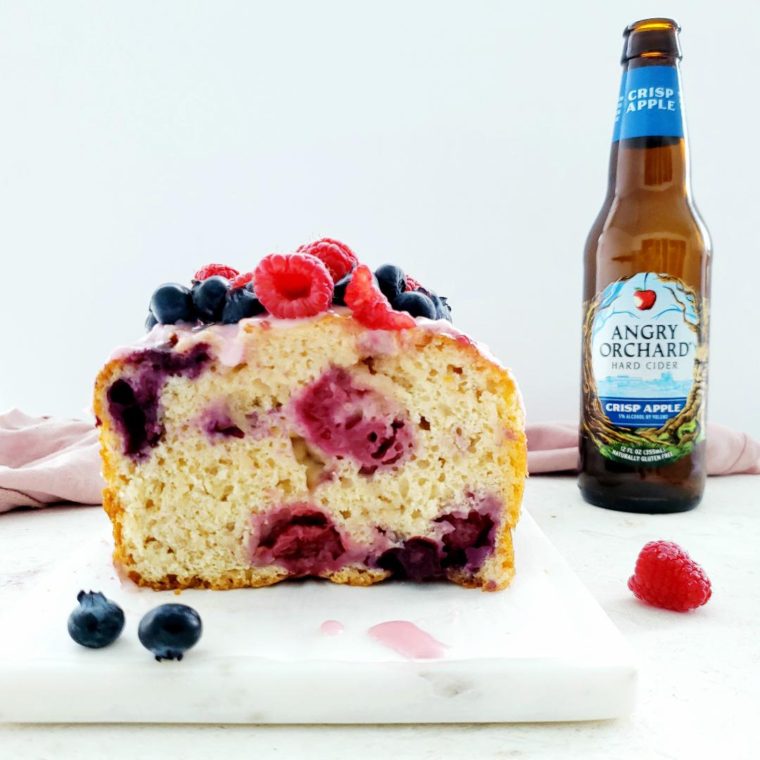 functional image hard apple cider beer berry bread blueberries raspberries side view of sliced loaf with blueberry and raspberry garnish on white marble cheeseboard back right is an angry orchard hard apple cider bottle