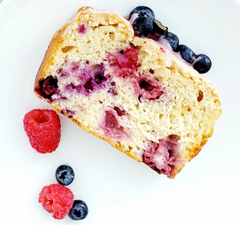 top down photo on white counter slice of hard apple cider bread so you can see the inside of the bread loaf is stuffed with baked in raspberries and blueberries top of loaf is iced with raspberry glaze and garnished with fresh blueberries photo is styled with two fresh raspberries and two fresh blueberries on the bottom left corner beer bread recipe
