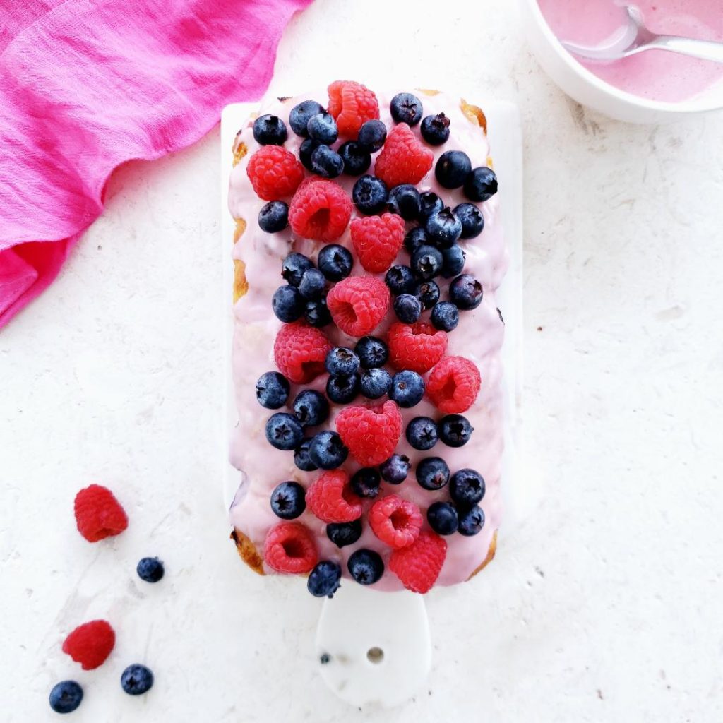 functional image apple cider bread top down photo on white limestone counter top of loaf is coated in a pink raspberry glaze then garnished with raspberries and blueberries upper right corner is a bowl of raspberry glaze with a silver spoon upper left is a pink ruffle linen beer bread recipe