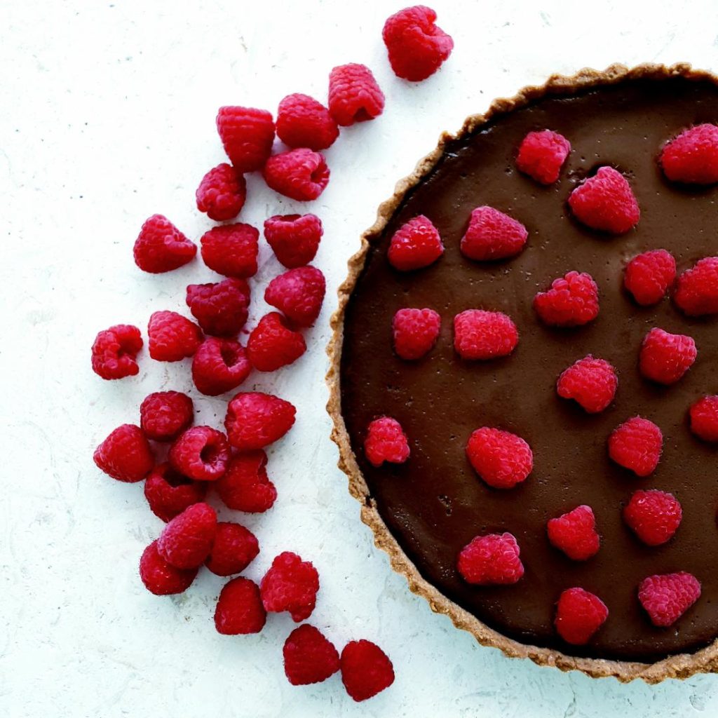 functional image raspberry chocolate tart top down view of uncut tart tart is on the left side of the photo the right side is styled with a lot of  loose raspberries