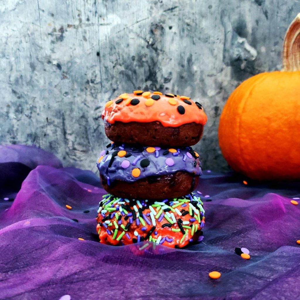 functional image halloweeen donuts  frosted chocolate fall donuts for fall decorated for halloween with orange and purple icing and different toppings stacked three high on purple tulle with a pumpkin in the right back corner
