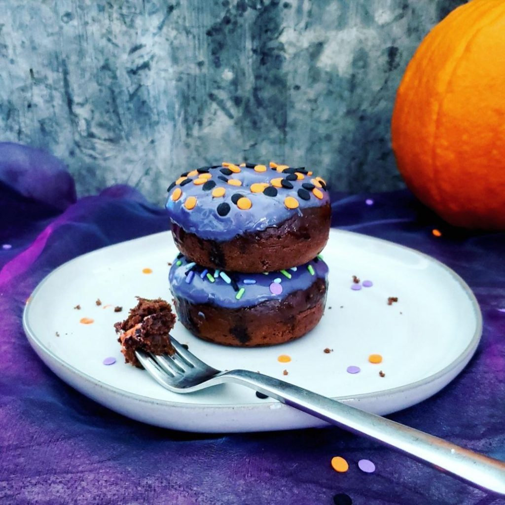 functional image halloween donuts  frosted chocolate fall donuts decorated for halloween with purple icing two donuts stacked on a light gray plate with a fork and halloween sprinkles