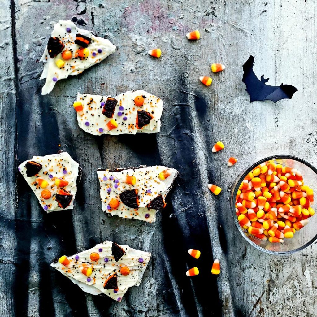 functional image white chocolate halloween oreo bark top down photo bark pieces arranged on a distressed board with spooky black and gray colors there is a bat in the upper left corner a bowl of candy corn on the middle right side and various pieces of candy corn scattered around bark
