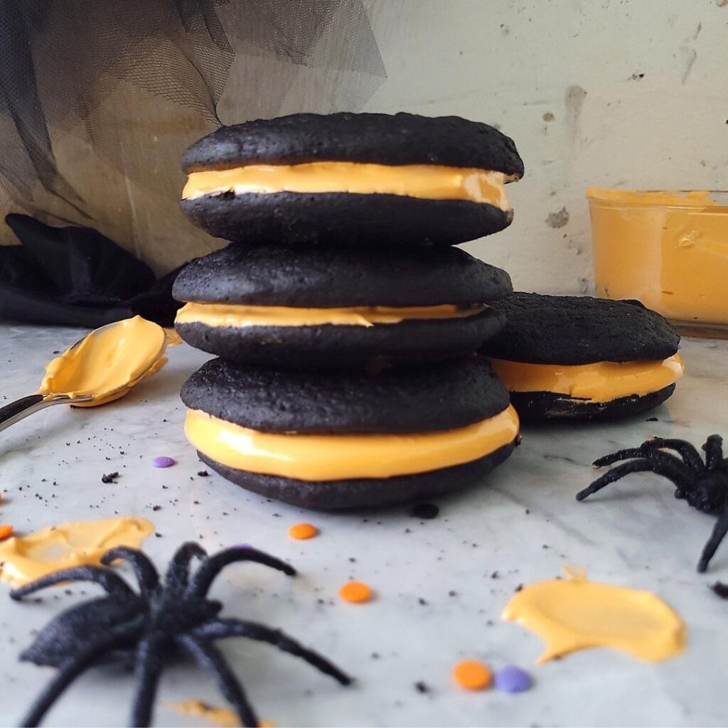 stack of dark chocolate halloween whoopie pies with orange filling. styled with spiders and halloween sprinkles.
