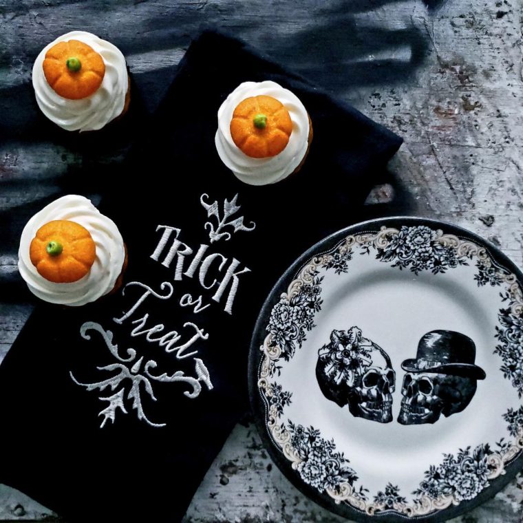 functional image pumpkin patch cupcakes top down photo cupcakes have white cream cheese icing piped on top with a glittery pumpkin shaped marshmallow three cupcakes total with a black trick or treat linen and a black and white plate with skulls for halloween