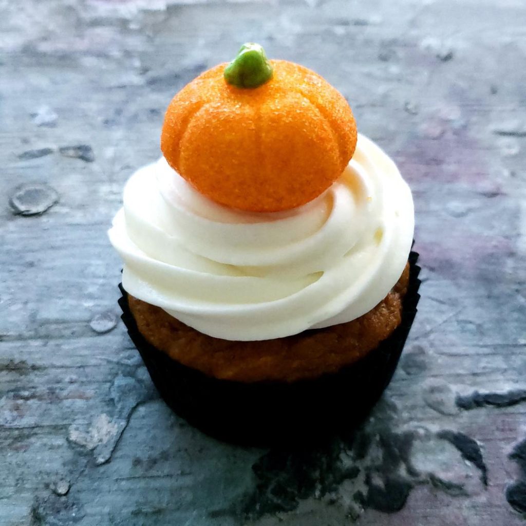 functional image pumpkin patch cupcake side view on distressed gray and purplish blue wood cupcake up close in a black cupcake liner with white cream cheese icing piped on and crowned with a glittery orange pumpkin shaped marshmallow